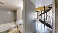 Located in the heart of Downtown Los Angeles on S Broadway, this two-story loft combines industrial character with modern finishes across two well-proportioned levels. The lower floor serves as the main living area, showcasing large-format polished concrete tile flooring, soaring ceilings, and oversized windows that draw in abundant natural light and urban views. A sleek, compact kitchen with white cabinetry and stainless steel appliances is tucked neatly along one wall, while a striking steel spiral staircase anchors the space as a sculptural focal point, connecting the two levels with an open mezzanine railing above. Exposed concrete beams and structural elements add raw, loft-style character throughout. The upper level hosts the generously sized primary bedroom, featuring polished tile floors, clean white walls, and access to the full bathroom. The bathroom is appointed with classic white subway tile, a soaking tub with shower, built-in open shelving, and a retro-patterned mosaic tile floor that adds a timeless design touch. A separate vanity area with a vessel sink and white cabinetry completes the bathroom. Throughout both floors, the interplay of industrial materials and clean, minimal finishes creates a cohesive and distinctive urban living environment.