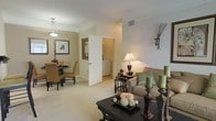 This single-floor residence offers a thoughtfully designed open-concept layout that seamlessly connects the living and dining areas, creating a cohesive and functional living environment. The space is finished in warm neutral tones throughout, complemented by plush wall-to-wall carpeting that adds comfort underfoot.

The living area is generously proportioned, providing ample room for relaxed seating arrangements. A stylish sofa with accent pillows anchors the space, accompanied by nesting side tables and a bold dark-finished coffee table, adding contrast against the light palette. Soft ambient lighting from a classic table lamp enhances the warm, inviting atmosphere.

Adjacent to the living area, the dining space accommodates a round dining table with upholstered chairs, ideal for intimate meals. Framed artwork adorns the walls, adding a curated, personal touch to the room. A pendant light fixture overhead provides focused illumination over the dining area.

A doorway leads to what appears to be a dedicated laundry area, offering the added convenience of in-unit washer and dryer access. Crown molding details and clean white door trim throughout contribute to the polished, finished appearance of the interior. The overall ambiance is calm, cohesive, and move-in ready.