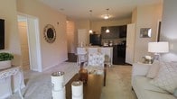 This single-floor residence is designed around an open-concept floor plan that seamlessly connects the living area to the kitchen, creating a cohesive and functional living environment. The living space features light-toned carpeting, neutral beige walls, and is accented by a decorative sunburst mirror that adds a stylish focal point to the room. A wooden coffee table with decorative accessories anchors the sitting area, while a patterned accent chair adds visual interest.

The kitchen is outfitted with dark cabinetry, a black refrigerator, and a peninsula-style breakfast bar with white bar stools, making it an inviting space for casual dining or entertaining. Pendant lights hang above the bar, providing warm, focused illumination. The countertops feature a stone-look finish that complements the darker cabinet tones.

Tile flooring in the kitchen and dining area transitions cleanly into the carpeted living space, subtly defining each zone. The overall ambiance is warm and welcoming, with consistent neutral tones throughout that lend the space a clean, move-in-ready aesthetic suited for comfortable everyday living.