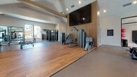 Located at 500 Lama Dr in Summerville, SC, this single-floor fitness center is a comprehensively outfitted facility designed to support a wide range of workout disciplines. The space is organized into distinct training zones, each thoughtfully appointed with purpose-built equipment and finishes.

The main strength training area features warm hardwood flooring, a large wall-mounted flat-screen TV set against a wood-paneled accent wall, and a selection of selectorized cable machines and weight benches. Full-length mirrors along one wall expand the sense of space and allow for form monitoring. A dedicated free weights section includes a dumbbell rack and ample open floor space for stretching or functional movements.

The cardio zone houses a row of modern treadmills positioned in front of large windows that frame outdoor views, flooding the area with natural light. Adjacent to this, stair climbers and air bikes occupy a spacious open area with wall-mounted displays.

A functional training zone is equipped with a ceiling-mounted rig system, TRX suspension trainers, kettlebells, medicine balls, and a heavy punching bag, catering to high-intensity and cross-training workouts. A separate, intimate cycling studio features two smart stationary bikes, flanked by low storage cabinetry and framed fitness artwork, with a large window offering views of palm trees outside. The overall ambiance is clean, modern, and motivating throughout.