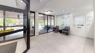 This well-designed building lobby offers a polished and welcoming entry experience spread across a single floor. The space is organized into distinct functional zones that flow seamlessly from one to the next. At the front, a bright and airy entrance lounge greets visitors with large floor-to-ceiling glass doors and windows that invite abundant natural light and offer views of the surrounding greenery. A decorative chandelier adds an elegant focal point overhead, while large-format light-toned porcelain tiles cover the floor. A seating area with contemporary grey upholstered armchairs and a side table provides a comfortable waiting space, partially enclosed by frosted glass partitions for a refined, semi-private feel. Moving deeper into the lobby, a textured accent wall with a wave-pattern finish adds visual interest alongside a built-in mailbox unit with individual metal compartments. The corridor transitions to grey carpet flooring, guiding residents toward the elevator bank. Recessed and track lighting throughout ensures consistent, well-distributed illumination across all areas. The overall design palette of crisp white walls, neutral tones, and modern finishes creates a clean, sophisticated ambiance that sets a professional tone for the entire building.