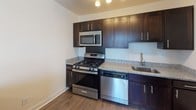 This single-floor unit presents a clean, neutral aesthetic throughout, with white walls and flush-mount ceiling lighting providing a bright, consistent ambiance across all rooms. The living area is generously proportioned, featuring wall-to-wall carpet and a large sliding glass door fitted with vertical blinds that allows abundant natural light to flow in. An additional window further enhances the airy feel of this space.

The kitchen is well-appointed with dark espresso cabinetry, granite-look countertops, and a full suite of stainless steel appliances including a gas range, built-in microwave, and dishwasher. Wood-look flooring grounds the kitchen, creating a practical contrast to the carpeted living spaces.

The bedrooms are comfortably sized, each featuring plush carpeting, ceiling-mounted lighting, and windows with blinds. Notably, one bedroom includes two open closets with hanging rods, offering generous storage capacity. A secondary bedroom provides a bright window view.

The bathroom is finished with large-format white marble-look tile surrounding a full tub-shower combination, complemented by brushed nickel fixtures and granite-look countertops, lending a clean and polished feel to the space.
