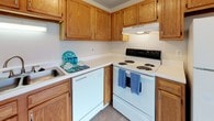 This single-floor residence offers a practical and comfortable layout across several well-defined spaces. The living area features neutral-toned carpet, a ceiling fan, and a window fitted with vertical blinds that allows natural light to filter in. A wall-mounted air conditioning unit provides climate control. An adjoining area with tile flooring creates a natural transition between spaces, with a second window adding additional brightness.

The kitchen is equipped with warm oak-toned cabinetry, white laminate countertops, a sink, a dishwasher, and a coil-burner electric range with an overhead exhaust hood. The layout makes efficient use of available space, with ample upper and lower cabinet storage.

The bedroom features plush carpeting, white walls, and a large window with vertical blinds that brings in natural light. A carpeted hallway connects the main areas and leads to the bathroom, which includes a single-sink vanity with wood cabinetry, tile flooring, and a full tub-shower combination with a curtain enclosure. A generously sized walk-in closet with built-in shelving and tile flooring provides substantial storage. The overall aesthetic is clean, neutral, and functional throughout.