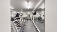 Located at 955 Broadway St in Boulder, Colorado, this fitness center offers a well-equipped workout space designed to accommodate a range of exercise needs. The room features a clean, utilitarian aesthetic with white walls, a textured ceiling, and durable dark rubber flooring that provides traction and cushioning throughout.

The cardio section includes a treadmill, an elliptical trainer, and a stationary spin bike, all positioned for easy access and use. For strength training, the space is outfitted with multiple racks of dumbbells spanning a variety of weights, as well as flat workout benches suitable for pressing and general conditioning exercises.

Large wall-mounted mirrors line a significant portion of the room, visually expanding the space and allowing users to monitor their form during workouts. A wall-mounted flat-screen television is positioned within clear sightlines of the cardio equipment, providing entertainment during sessions. Bright fluorescent overhead lighting ensures the entire space is well illuminated.

The layout is efficient, making practical use of the available footprint while keeping equipment organized and accessible. The overall ambiance is functional and purposeful, reflecting a straightforward approach to fitness facility design suited to the needs of its users in the heart of Boulder.