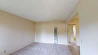 This single-floor space presents a clean, minimalist interior characterized by neutral beige walls and light-colored carpeting throughout. The main room is a generously sized, open area with smooth white ceilings that contribute to a bright and airy atmosphere. A wall-mounted vertical HVAC unit is centrally positioned along the main wall, providing both heating and cooling functionality directly within the room. The walls are finished in a warm off-white tone, complementing the soft, textured carpeting underfoot. An open doorway on the right side of the room leads to an adjacent space, suggesting additional rooms beyond. The overall layout is straightforward and functional, with no built-in shelving or decorative architectural detailing. The space is currently vacant, allowing for a clear view of its structural elements. The lighting appears to be natural and ambient, contributing to the room's clean and understated aesthetic. The simplicity of the design makes it highly adaptable for various uses.