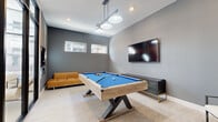 This contemporary game room is thoughtfully designed as a dedicated recreational space, offering an inviting and energetic atmosphere for social gatherings. At the heart of the room sits a striking pool table featuring a rustic wood frame with bold cross-leg bases and vivid blue felt, immediately drawing the eye upon entry. Light-toned large-format tile flooring extends throughout, lending a clean, polished foundation to the space.

The room is painted in sophisticated shades of gray, creating a modern, sleek aesthetic. A stylish multi-pendant light fixture hangs directly above the pool table, providing focused illumination, while recessed ceiling lights ensure the entire space is well-lit. A wall-mounted flat-screen television is positioned prominently on one wall, making the room equally suited for viewing entertainment alongside gameplay. A cue rack is mounted nearby for convenient access.

Seating is thoughtfully arranged around the perimeter, including a cognac leather sofa, additional lounge chairs, and a decorative oval ottoman, offering comfortable spots for spectators. Horizontal accent windows along one wall allow natural light to filter in, while a full-height glass partition wall with a black steel frame opens the room visually to an adjacent bright lounge area, enhancing the sense of openness and connectivity throughout the overall space.
