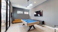 This contemporary game room is thoughtfully designed as a dedicated recreational space, offering an inviting and energetic atmosphere for social gatherings. At the heart of the room sits a striking pool table featuring a rustic wood frame with bold cross-leg bases and vivid blue felt, immediately drawing the eye upon entry. Light-toned large-format tile flooring extends throughout, lending a clean, polished foundation to the space.

The room is painted in sophisticated shades of gray, creating a modern, sleek aesthetic. A stylish multi-pendant light fixture hangs directly above the pool table, providing focused illumination, while recessed ceiling lights ensure the entire space is well-lit. A wall-mounted flat-screen television is positioned prominently on one wall, making the room equally suited for viewing entertainment alongside gameplay. A cue rack is mounted nearby for convenient access.

Seating is thoughtfully arranged around the perimeter, including a cognac leather sofa, additional lounge chairs, and a decorative oval ottoman, offering comfortable spots for spectators. Horizontal accent windows along one wall allow natural light to filter in, while a full-height glass partition wall with a black steel frame opens the room visually to an adjacent bright lounge area, enhancing the sense of openness and connectivity throughout the overall space.