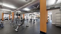 Located at 3227 S Parker Rd in Denver, Colorado, this fitness facility offers a generously sized, single-floor exercise space designed to accommodate a wide range of workout needs. The open-concept layout provides ample room for both cardio and strength training, with distinct zones clearly defined throughout the floor plan.

The cardio area features a row of treadmills and elliptical machines positioned along large windows, allowing natural light to fill the space and providing views to the outside. Full-length mirrors line multiple walls, enhancing the sense of openness and allowing users to monitor their form throughout their workouts.

The strength training zone is well-equipped with cable machines, weight racks, barbells, and bench press stations, offering comprehensive resistance training options. A dedicated cycling area houses multiple stationary bikes beneath ceiling-mounted fans and flatscreen TVs, creating an engaging indoor cycling environment.

Architectural details include exposed ductwork, structural columns painted in a bold orange accent, white brick walls, and a ceiling with distinctive ribbed paneling. Durable rubber flooring covers the entire space, and industrial-style pendant and linear lighting ensures the area is well-lit throughout. The combination of functional design and modern finishes creates a professional and motivating atmosphere suited for all fitness levels.
