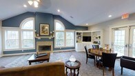 This single-floor living space makes a strong first impression with its vaulted ceiling that rises dramatically above the main living area, creating an open and airy atmosphere. The walls are painted in a rich slate blue, providing a bold yet sophisticated backdrop throughout the room. A tile-surround fireplace serves as the focal point of one wall, flanked by white trim and framed artwork, adding both warmth and visual interest. Above the fireplace, a large arched window floods the space with natural light, complementing the additional standard windows nearby that offer views of the surrounding greenery. The flooring features a patterned area rug layered over carpet, defining the seating area, which includes a tan leather sofa and a round accent table. A built-in corner cabinetry unit with white shelving, display space, and lower cabinets houses a flat-screen television, providing practical storage while maintaining a clean, cohesive look. Adjacent to the built-ins, a round dark-wood dining table with upholstered chairs offers a dedicated space for dining or casual gatherings. Recessed lighting and a ceiling fan ensure the space is well-lit and comfortable year-round.