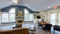This single-floor living space makes a strong first impression with its vaulted ceiling that rises dramatically above the main living area, creating an open and airy atmosphere. The walls are painted in a rich slate blue, providing a bold yet sophisticated backdrop throughout the room. A tile-surround fireplace serves as the focal point of one wall, flanked by white trim and framed artwork, adding both warmth and visual interest. Above the fireplace, a large arched window floods the space with natural light, complementing the additional standard windows nearby that offer views of the surrounding greenery. The flooring features a patterned area rug layered over carpet, defining the seating area, which includes a tan leather sofa and a round accent table. A built-in corner cabinetry unit with white shelving, display space, and lower cabinets houses a flat-screen television, providing practical storage while maintaining a clean, cohesive look. Adjacent to the built-ins, a round dark-wood dining table with upholstered chairs offers a dedicated space for dining or casual gatherings. Recessed lighting and a ceiling fan ensure the space is well-lit and comfortable year-round.