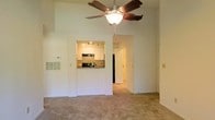 Located at 42 Magnolia, this single-floor residence makes an immediate impression with its notably high ceilings, which lend an airy, open feel to the main living area. A wood-bladed ceiling fan with a central light fixture is mounted overhead, providing both circulation and ambient lighting while emphasizing the generous vertical space above.

The living area features neutral-toned wall-to-wall carpeting that extends throughout, creating a cohesive and comfortable foundation underfoot. A pass-through opening connects the living space to the kitchen, where white cabinetry lines the walls beneath track lighting, offering ample storage. Built-in appliances, including an oven and what appears to be a refrigerator, are integrated into the kitchen layout, and a countertop is visible at the pass-through window.

A hallway visible from the main living area leads further into the unit, suggesting additional rooms beyond. The overall color palette is light and neutral throughout, with white walls and trim contributing to a clean, bright ambiance. The layout flows naturally from the main living space toward the kitchen and adjoining areas, offering a functional and well-connected floor plan.