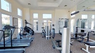This fitness center offers a comprehensive workout environment designed to accommodate a variety of exercise routines. The space is characterized by soaring ceilings with a recessed tray detail, lending an open and expansive feel throughout. Large transom windows and glass-paneled doors line the walls, flooding the interior with natural light and providing views of the surrounding exterior, creating an inviting atmosphere for exercise.

The flooring features a durable, textured carpet well-suited for a high-traffic fitness environment. The layout is thoughtfully organized to maximize usable space while allowing comfortable movement between equipment stations.

The cardio section includes a StairMaster step climber and a stationary cycling bike, ideal for cardiovascular conditioning. The strength training area is equipped with multiple cable and resistance machines, including a multi-functional cable station and a seated strength unit, catering to both upper and lower body workouts. Reference charts posted on the walls provide guidance on proper equipment use.

The bright, neutral color palette of white and cream walls enhances the sense of openness, while recessed lighting supplements the generous natural light. Overall, this fitness center presents a clean, well-maintained, and fully functional space suitable for a broad range of fitness needs.