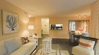 This single-story space presents an open-concept layout that seamlessly connects the living and dining areas, creating a cohesive and functional flow throughout. The living area is finished with warm peach-toned walls that lend a welcoming ambiance, complemented by dark hardwood flooring that runs consistently through the space. A built-in wall shelf unit serves as a practical and decorative focal point, offering display space between the living and dining zones. The dining area, positioned near a window that allows natural light to filter in, accommodates a classic dining table and upholstered seating. A hallway visible from the main living area leads to additional rooms, suggesting a well-organized floor plan. The living space is generously proportioned, accommodating a sofa with accent pillows, a side table with a lamp, and a central coffee table atop a light area rug. Ceiling-mounted light fixtures provide consistent overhead illumination throughout. White baseboards and trim provide clean contrast against the darker flooring, while a visible HVAC vent and thermostat reflect practical, functional design considerations integrated throughout the space.