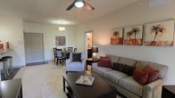 This single-floor residence offers a comfortable and well-appointed living environment designed with both style and functionality in mind. The open-concept layout seamlessly connects the living and dining areas, creating a spacious and sociable atmosphere throughout.

The living area is anchored by a plush gray sofa accented with warm red and brown throw pillows, paired with a patterned accent chair and a dark wood coffee table. Decorative wall art featuring tropical motifs adds a touch of warmth and personality to the neutral-toned walls. A ceiling fan with a light fixture overhead provides both comfort and illumination, keeping the space bright and airy.

Just beyond the living area, the dining space accommodates a dark wood table and cushioned chairs beneath a pendant light fixture, making it an inviting spot for meals and gatherings. The consistent use of warm neutrals and carpeted flooring throughout ties the two areas together cohesively.

A hallway visible from the main living space suggests additional rooms beyond, hinting at a thoughtfully laid-out floor plan. The overall ambiance is cozy yet refined, with carefully chosen furnishings and decor that create a move-in-ready aesthetic suitable for everyday living.
