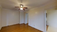 This single-floor space showcases timeless architectural character through its prominent crown molding, which frames the ceiling and adds a refined touch to the interior. The room is finished with warm hardwood flooring that runs throughout, providing both durability and visual continuity. A multi-blade ceiling fan with an integrated light fixture serves as the central overhead lighting source, offering both illumination and air circulation. The walls are painted in a neutral off-white tone, creating a bright and versatile backdrop suitable for a variety of uses. Crisp white baseboards complement the crown molding, reinforcing the traditional detailing found throughout the space. A white-paneled interior door provides access to an adjoining area, suggesting a connected layout. The overall ambiance is clean, understated, and move-in ready, with classic design elements that lend the space a sense of character and warmth. The combination of natural wood tones and neutral wall finishes creates a cohesive, inviting atmosphere well-suited for comfortable everyday living.