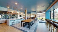 Located at 2337 S Blackhawk St in Aurora, Colorado, this contemporary single-floor community clubhouse is thoughtfully designed to foster social interaction and comfort. The space boasts a striking interior palette of teal-painted ceilings, light wood-plank flooring, and mixed tile accents that create a vibrant yet cohesive atmosphere throughout.

At the heart of the space is a stylish bar area clad in light blue tile, complete with pendant lighting suspended from copper-toned rods and a mounted flat-screen TV. A large communal table with woven stools anchors the central gathering area, ideal for collaborative use. Adjacent to the bar, a full communal kitchen features sleek wood cabinetry, a white countertop island, and a dramatic wall of dark hexagonal tiles with open shelving stocked with colorful dishware.

Multiple seating zones provide variety for visitors—a cozy nook with upholstered brown leather and gray banquette seating, decorative pillows, and pegboard shelving offers an intimate retreat, while a window-side bar counter lined with modern high-back stools delivers unobstructed views of the surrounding green landscape. Floor-to-ceiling windows and expansive glass doors flood the interior with natural light, connecting the indoors seamlessly with the outdoors. An exposed brick accent wall adds warmth and texture to complete the space's modern-industrial character.