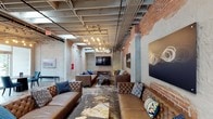 Located at 1400 S 2nd St in Wilmington, NC, this single-floor space blends raw industrial character with modern design sensibility. The interior showcases exposed brick walls, structural concrete columns, and an open ceiling with visible ductwork and conduit — all signature elements of an industrial-chic aesthetic. Pendant Edison bulb chandeliers and track lighting cast a warm, inviting glow throughout the space, enhancing its urban atmosphere.

The main lounge area is generously proportioned, offering a spacious and open layout ideal for gatherings or events. Tufted brown leather sofas and armchairs are arranged in conversational groupings atop artistically patterned area rugs, providing both comfort and visual interest. Colorful accent pillows add pops of blue and teal against the rich leather tones.

A wall-mounted flat-screen display serves as a focal point at the far end of the room, making the space well-suited for presentations or entertainment. Curated wall art, including abstract sculptural pieces and bold canvas works, contributes to the space's contemporary character. Natural light filters in through windows, complementing the ambient lighting and highlighting the textured surfaces of brick and concrete throughout.
