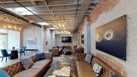 Located at 1400 S 2nd St in Wilmington, NC, this single-floor space blends raw industrial character with modern design sensibility. The interior showcases exposed brick walls, structural concrete columns, and an open ceiling with visible ductwork and conduit — all signature elements of an industrial-chic aesthetic. Pendant Edison bulb chandeliers and track lighting cast a warm, inviting glow throughout the space, enhancing its urban atmosphere.

The main lounge area is generously proportioned, offering a spacious and open layout ideal for gatherings or events. Tufted brown leather sofas and armchairs are arranged in conversational groupings atop artistically patterned area rugs, providing both comfort and visual interest. Colorful accent pillows add pops of blue and teal against the rich leather tones.

A wall-mounted flat-screen display serves as a focal point at the far end of the room, making the space well-suited for presentations or entertainment. Curated wall art, including abstract sculptural pieces and bold canvas works, contributes to the space's contemporary character. Natural light filters in through windows, complementing the ambient lighting and highlighting the textured surfaces of brick and concrete throughout.