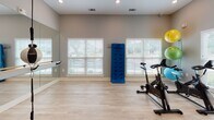 Located at 10 Shell Creek Dr in Port Royal, South Carolina, this dedicated fitness studio is thoughtfully designed to support a wide range of workout routines. The single-floor space features light hardwood-style flooring that contributes to a clean, open feel, making it well-suited for cardio, strength, and flexibility training alike.

A full-length mirrored wall runs along one side of the room, paired with a classic ballet barre — ideal for stretching, barre workouts, or dance-based exercise. Opposite the mirrors, multiple windows with white blinds allow generous natural light to fill the space, creating an inviting and energizing atmosphere. A ceiling fan with recessed lighting ensures comfortable airflow during intense sessions.

The studio is equipped with spin bikes for cardiovascular training, a CrossBody Trainer freestanding punching system for boxing and functional fitness, and a wall-mounted rack of medicine balls for strength and conditioning exercises. A large flat-screen television is mounted on the wall, perfect for streaming workout classes or instructional content. A yoga/exercise mat is also present for floor-based exercises.

The space connects directly to an adjacent area through a glass-paned door, suggesting it is part of a larger facility. Overall, this fitness studio combines practical equipment, smart layout, and a bright, motivating environment to deliver a comprehensive at-home or community workout experience.