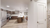 This thoughtfully designed 2-bedroom, 1-bathroom residence delivers a functional and stylish single-floor layout. The open-concept kitchen and living area serves as the heart of the home, featuring rich medium-toned wood cabinetry, a white subway tile backsplash, stainless steel appliances, and a central island with a built-in sink and bar seating — ideal for both meal prep and casual dining. Floating wood shelves add an airy, contemporary touch, while light wood-style flooring runs throughout, lending warmth and visual continuity. A sliding barn door adds a charming architectural detail to the shared living and dining space, which is brightened by multiple windows. The living room offers a comfortable, light-filled retreat with natural light filtering through large windows with views to the exterior. The spacious primary bedroom includes a wall-mounted mini-split system for personalized climate control and a generous window for natural light. The second bedroom is equally well-appointed. The full bathroom features a tiled walk-in shower, white vanity, and clean, minimalist finishes. A dedicated in-unit laundry room with side-by-side Whirlpool washer and dryer provides added convenience. Two closets offer ample storage throughout.