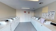 This shared laundry facility is a single-room space designed for efficient, communal use. The room is laid out with rows of top-loading washing machines and dryers lining both sides, providing ample capacity for residents. The machines are white and well-maintained, each equipped with coin or card-operated payment systems managed through a CSC service network. Detergent dispensers are available on the machines for added convenience.

The interior features light gray flooring, neutral-toned walls, and white vertical paneling on one end wall, contributing to a clean and functional aesthetic. Overhead fluorescent lighting ensures the space is well-lit throughout. Ventilation is provided by ceiling vents positioned throughout the room.

A bright red entry door with glass pane inserts is located at one end of the room, allowing natural light to filter in and providing a clear view to the exterior. A fire extinguisher is mounted on the wall, and informational postings — including laundry tips, drying instructions, and service contact information — are displayed for user reference. The overall ambiance is utilitarian and orderly, prioritizing ease of use and accessibility for all users.
