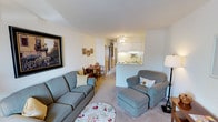 This single-floor residence offers a well-organized and comfortable layout designed for easy, practical living. The living room is carpeted and generously proportioned, accommodating a full sofa, armchair with ottoman, and a decorative area rug. Neutral walls and soft lighting create a warm, inviting ambiance, complemented by a window dressed with sheer curtains that allows natural light to filter through. A framed artwork piece anchors one wall, adding a touch of character to the space.

The kitchen is semi-open, visible from the living area via a pass-through counter, and is outfitted with white cabinetry, a coil-burner electric range, a standard refrigerator, a stainless steel sink, and white tile backsplash. A ceiling fan with a light fixture provides ventilation overhead. Adjacent to the kitchen, a small dining area comfortably seats a compact table and chairs.

The bedroom is a calm, softly lit retreat featuring carpet flooring, a window with curtains, and built-in wooden shelving and closet storage that offers ample organization. The bathroom includes a tiled walk-in shower with a sliding glass door, complemented by clean, light-toned finishes throughout.