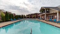 Located at 4401 E 46th St in North Little Rock, Arkansas, this outdoor amenity space centers around a generously sized resort-style swimming pool with striking blue tile accents and crystal-clear water. The pool deck is finished in a warm brick-toned surround and lined with an abundance of blue lounge chairs, offering ample space for relaxation and sunbathing. Elegant white columns frame the perimeter, adding a classic architectural touch that complements the surrounding multi-story brick buildings with pitched slate-gray rooflines. A covered pavilion constructed of matching brick anchors one side of the space, providing a shaded outdoor gathering area equipped with grills and seating — ideal for entertaining. Tall, neatly trimmed evergreen trees are arranged along the pool's edge, creating natural privacy screens and adding lush greenery to the environment. The clubhouse building, accessible directly from the pool deck, features large windows, a covered balcony with decorative iron railings, and white columned entries that reinforce the upscale character of the community. The overall ambiance is that of a well-appointed, resort-inspired outdoor retreat designed for both leisure and social gatherings.