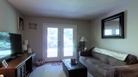 This single-floor space offers a comfortable and functional layout designed for relaxed everyday living. The living area serves as the heart of the home, highlighted by a pair of French doors that flood the room with natural light and frame a lush, tree-lined outdoor view, creating a seamless connection between the interior and the surrounding greenery. Plush carpeting throughout the living area adds warmth and comfort underfoot, complementing the neutral, earthy tones of the walls and furnishings. A textured ceiling adds a subtle architectural detail to the space. The room is furnished with a generous sofa and a leather ottoman that doubles as a coffee table, offering both style and practicality. A classic table lamp on a side table provides warm ambient lighting, while a decorative mirror on the wall enhances the sense of space and depth. An entertainment console along one wall provides additional storage and functionality. The overall ambiance of the space is cozy and welcoming, with an emphasis on natural light and a calm, neutral color palette that makes the interior feel open and comfortable throughout.