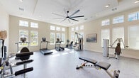 Located in Wesley Chapel, FL, this dedicated fitness center offers a spacious, single-floor exercise room designed to support a complete workout experience. The room features soaring ceilings that amplify the sense of openness, complemented by an oversized multi-blade ceiling fan that ensures comfortable air circulation throughout. Large windows along multiple walls, including elevated transom windows, flood the space with abundant natural light and provide views of the surrounding community. The light-toned walls and speckled fitness flooring create a clean, welcoming atmosphere. A diverse array of equipment is thoughtfully arranged throughout, including treadmills, elliptical trainers, a recumbent bike, cable machines, a multi-purpose weight bench, and a rack of dumbbells. A large wall-mounted mirror and an accent wall in warm amber highlight a dedicated strength training zone, while built-in open shelving adds functional storage. Recessed lighting supplements the natural light for evening use. Decorative wall art and tasteful accents contribute to an inviting ambiance that balances functionality with aesthetic appeal. Additional doors lead to adjoining utility or support spaces, rounding out this well-appointed community fitness facility.