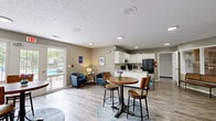 Located at 8016 E Louisville Dr in Lawrence, Indiana, this single-floor community space seamlessly combines a fully equipped kitchen with a welcoming dining and lounge area. The kitchen is outfitted with white cabinetry, granite-style countertops, a double-basin sink set within a central peninsula, black appliances including a refrigerator, electric range, and microwave, providing a practical and well-appointed setup for communal use. Light wood-look flooring runs throughout, complementing the neutral gray walls and textured white ceiling, creating a clean and cohesive aesthetic. The open-plan dining area accommodates multiple bistro-style round tables with bar seating, offering a casual and social atmosphere. Large windows dressed with sheer curtains and a set of glass French doors bathe the space in natural light and provide direct views and access to an outdoor pool area, enhancing the sense of openness. A seating nook with a pair of blue armchairs adds a comfortable lounge corner, while a oversized Connect Four game contributes a playful, recreational element to the space. Recessed and flush-mount ceiling lights ensure bright, even illumination throughout. This thoughtfully designed community space balances functionality with a relaxed, inviting ambiance suited for dining, socializing, and leisure activities.