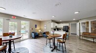 Located at 8016 E Louisville Dr in Lawrence, Indiana, this single-floor community space seamlessly combines a fully equipped kitchen with a welcoming dining and lounge area. The kitchen is outfitted with white cabinetry, granite-style countertops, a double-basin sink set within a central peninsula, black appliances including a refrigerator, electric range, and microwave, providing a practical and well-appointed setup for communal use. Light wood-look flooring runs throughout, complementing the neutral gray walls and textured white ceiling, creating a clean and cohesive aesthetic. The open-plan dining area accommodates multiple bistro-style round tables with bar seating, offering a casual and social atmosphere. Large windows dressed with sheer curtains and a set of glass French doors bathe the space in natural light and provide direct views and access to an outdoor pool area, enhancing the sense of openness. A seating nook with a pair of blue armchairs adds a comfortable lounge corner, while a oversized Connect Four game contributes a playful, recreational element to the space. Recessed and flush-mount ceiling lights ensure bright, even illumination throughout. This thoughtfully designed community space balances functionality with a relaxed, inviting ambiance suited for dining, socializing, and leisure activities.