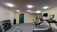 This single-floor fitness room provides a well-equipped exercise environment designed for convenient, everyday use. The space features neutral beige walls and grey carpet flooring, creating a clean and functional atmosphere. Overhead, multiple flush-mount ceiling lights ensure consistent, bright illumination throughout the room. A wall-mounted flat-screen television is positioned to be visible from the cardio equipment, enhancing the workout experience. The room is outfitted with a selection of cardio machines, including an elliptical trainer, a stationary bike, and a treadmill from the True Fitness brand. A weight bench and additional exercise equipment are also present, offering options for strength training. A water cooler station is conveniently located near the entrance, supporting hydration during workouts. The room is accessed through a sturdy steel door with a small window, and ventilation is provided via ceiling-mounted HVAC vents. The overall layout is open and straightforward, maximizing usable floor space while keeping the environment practical and easy to navigate.