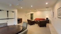 This single-floor space functions as a well-appointed lounge or common area, designed to accommodate both social gatherings and relaxed conversation. The open layout is finished with neutral-toned walls and light carpeting throughout, creating a clean and inviting atmosphere. Recessed lighting is distributed evenly across the ceiling, providing consistent, warm illumination across the entire room.

The seating area serves as the focal point of the space, anchored by a curved burnt-orange sofa paired with two brown upholstered armchairs arranged around a low coffee table. This arrangement encourages informal conversation and comfortable lounging. Additional seating is available at a small round table near the foreground, accompanied by cushioned chairs, offering a more intimate setting for smaller groups.

Along one wall, a built-in cabinetry unit houses a beverage refrigerator, adding a functional hospitality element to the space. A doorway visible toward the back of the room suggests access to an adjacent area or corridor, contributing to the overall flow of the layout. The combination of practical amenities and comfortable furnishings makes this space well-suited for use as a community lounge, hospitality suite, or collaborative gathering area.