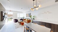 Located at 333 15th St NE in Washington, DC's Capitol Hill neighborhood, this community amenity space at Viva Capitol Hill offers a thoughtfully designed environment for residents to gather and relax. The interior is characterized by crisp white walls, large-format tile flooring, and recessed lighting, creating a bright and welcoming atmosphere throughout.

The main interior area is divided into two functional zones. A dining area anchored by a rectangular table and mid-century modern wood chairs sits beneath a striking brass and globe pendant light fixture, adding warmth and a contemporary flair. Curated artwork lines the corridor walls, lending a gallery-like quality to the space. Adjacent to the dining zone, a comfortable lounge area features a cognac leather sofa, deep plum swivel chairs, a glass-topped coffee table, and a terrazzo-patterned rug. Large windows allow natural light to fill the seating area, and a low dark-wood credenza with a white marble countertop and utility sink serves as a functional built-in along one wall.

An accessible restroom with dark tile flooring and ADA-compliant fixtures is also included. The space opens to a multi-level outdoor courtyard with brick pavers, string lighting, a gas grill, and lush greenery—ideal for outdoor entertaining. The building's modern exterior features grey panel cladding, steel railings, and expansive windows.
