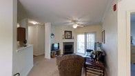 This single-floor space centers around a comfortable living area designed with both function and warmth in mind. The room features a corner fireplace with a dark tile surround and a classic white mantle, serving as a natural focal point. Crown molding lines the ceiling, adding a refined architectural detail, while a ceiling fan with a light fixture provides both airflow and ambient lighting. Soft, neutral-toned carpet covers the floor throughout, contributing to the room's cozy feel. A sliding glass door allows natural light to filter in through sheer curtains, offering a view of the outdoors and providing easy access to an exterior space. Structural columns frame the living area, subtly defining the layout while maintaining an open feel. A media setup with a television and media console is positioned near the fireplace, making the space well-suited for everyday living and entertainment. The overall palette of warm beige and taupe tones, combined with traditional furnishings and architectural details, gives this space a classic, comfortable residential character.