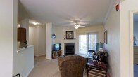 This single-floor space centers around a comfortable living area designed with both function and warmth in mind. The room features a corner fireplace with a dark tile surround and a classic white mantle, serving as a natural focal point. Crown molding lines the ceiling, adding a refined architectural detail, while a ceiling fan with a light fixture provides both airflow and ambient lighting. Soft, neutral-toned carpet covers the floor throughout, contributing to the room's cozy feel. A sliding glass door allows natural light to filter in through sheer curtains, offering a view of the outdoors and providing easy access to an exterior space. Structural columns frame the living area, subtly defining the layout while maintaining an open feel. A media setup with a television and media console is positioned near the fireplace, making the space well-suited for everyday living and entertainment. The overall palette of warm beige and taupe tones, combined with traditional furnishings and architectural details, gives this space a classic, comfortable residential character.
