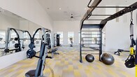Located at 16630 W Pat Tillman Blvd in Surprise, AZ, this fitness center offers a spacious, single-floor layout thoughtfully designed to accommodate a full range of workout activities. The facility is characterized by soaring ceilings that create an open, airy atmosphere, complemented by large windows positioned along multiple walls that flood the space with natural light and provide views of the surrounding exterior. A ceiling fan overhead aids in ventilation and comfort throughout the workout area.

The flooring features a striking geometric-patterned carpet in yellow, gray, and white tones, adding a modern, energetic aesthetic to the environment. A full-length wall mirror on one side visually expands the space while allowing users to monitor their form during exercise.

The fitness center is comprehensively equipped with a dedicated cardio zone housing treadmills, ellipticals, and stationary cycling bikes — including smart-enabled models with integrated screens. Wall-mounted flat-screen televisions provide entertainment during cardio sessions. A strength training area includes a multi-station cable machine, a functional training rig with pull-up bars and ladder attachments, free weights, benches, and medicine balls. An exercise mat area supports stretching and floor-based workouts. The layout provides ample open floor space, ensuring comfortable movement between equipment stations throughout the facility.