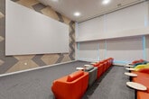 Located at 800 Harbor Blvd in Weehawken, NJ, this screening and lounge room is a thoughtfully designed amenity space that blends entertainment functionality with modern, energetic aesthetics. The main room is generously proportioned, featuring soaring ceilings that amplify the sense of openness and a large projection screen anchored to a striking feature wall adorned with a bold chevron pattern in warm beige and grey tones. The flooring throughout is a neutral grey carpet that grounds the vibrant furnishings above. Rows of curved, low-profile lounge chairs in a lively mix of orange, yellow, teal, and grey are arranged in a tiered, theater-style layout, each accompanied by compact round side tables for added convenience. Along the perimeter, tall windows fitted with roller shades allow controlled natural light to filter in, accented by subtle blue LED lighting along the window sills that adds a contemporary, cinematic ambiance. A structural column punctuates the center of the room, seamlessly integrated into the layout. The space is accessed through a well-appointed entry foyer featuring textured grey walls, recessed linear lighting, and a sleek floating wood credenza with a dark countertop — providing a polished and functional transition into the main room.