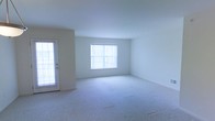 This single-floor space presents a clean, minimalist layout designed to maximize both comfort and functionality. The main living area is generously proportioned, featuring soft, light-colored wall-to-wall carpeting that adds warmth underfoot and contributes to the overall sense of openness. Crisp white walls provide a neutral backdrop throughout, making the space feel bright and airy.

Natural light is a defining feature of this space. A large window dressed with vertical blinds dominates one wall of the main living area, allowing ample daylight to pour in. A glass-paneled exterior door on the adjacent wall further enhances the brightness and provides convenient access to the outdoors.

The layout is straightforward and versatile, lending itself well to a variety of furniture arrangements and uses. The open floor plan ensures a smooth flow between areas, while the consistent flooring and wall finishes create a cohesive, unified aesthetic throughout. The overall ambiance is calm and understated, offering a blank canvas that can be easily personalized to suit a wide range of needs and preferences. This well-lit, thoughtfully designed space combines practical features with a welcoming atmosphere.