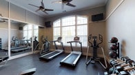This single-floor fitness room offers a focused and functional workout environment designed to support cardiovascular training. The space is anchored by a row of cardio equipment, including two treadmills and an elliptical machine positioned near the front, with additional equipment visible along the mirrored wall to the left. The full-length mirror running along one wall visually expands the room while allowing users to monitor their form during exercise.

Natural light filters generously into the space through a large arched window with white trim, lending an open, airy quality to the interior. The warm, neutral-toned walls complement the dark rubber flooring, which provides a durable and slip-resistant surface suited for high-activity use. Two ceiling fans are mounted overhead, promoting air circulation and comfort during intense workouts.

Two wall-mounted flat-screen televisions are positioned at viewing angles accessible from the cardio equipment, allowing users to stay entertained during sessions. Crown molding along the upper walls adds a refined architectural touch. Overall, the space balances practicality with a clean, polished aesthetic.