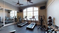This single-floor fitness room offers a focused and functional workout environment designed to support cardiovascular training. The space is anchored by a row of cardio equipment, including two treadmills and an elliptical machine positioned near the front, with additional equipment visible along the mirrored wall to the left. The full-length mirror running along one wall visually expands the room while allowing users to monitor their form during exercise.

Natural light filters generously into the space through a large arched window with white trim, lending an open, airy quality to the interior. The warm, neutral-toned walls complement the dark rubber flooring, which provides a durable and slip-resistant surface suited for high-activity use. Two ceiling fans are mounted overhead, promoting air circulation and comfort during intense workouts.

Two wall-mounted flat-screen televisions are positioned at viewing angles accessible from the cardio equipment, allowing users to stay entertained during sessions. Crown molding along the upper walls adds a refined architectural touch. Overall, the space balances practicality with a clean, polished aesthetic.