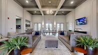 This refined single-floor space makes a striking first impression with its dramatic, soaring coffered ceilings adorned with decorative tin-style inlays and anchored by a statement chandelier. Tall white columns and expansive floor-to-ceiling walls define the open, airy atmosphere of the combined living and dining area, which serves as the heart of the space. The living area is generously appointed with plush velvet sofas, upholstered ottomans, and layered area rugs over large-format light tile flooring, creating a welcoming and polished environment. A wall-mounted flat-screen television adds a functional focal point to one end of the room. The dining area features a substantial dark wood communal table surrounded by neutral upholstered chairs, with elegant wall sconces providing warm accent lighting. Multiple sets of glass French doors flood the space with natural light and offer views of an outdoor terrace. A private office off the hallway is carpeted and bathed in natural light from oversized windows dressed with sheer curtains, and is furnished with a functional executive desk setup — ideal for professional consultations. Lush potted plants and curated wall art throughout add a fresh, sophisticated character to the overall ambiance.