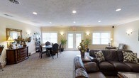 This single-floor space functions as a combined lounge and dining area designed for comfort and casual gathering. The room features a textured ceiling with recessed lighting, which provides even, ambient illumination throughout. Warm beige walls contribute to an inviting atmosphere, complemented by wall sconces that add a soft, decorative glow on either side of the central French doors.

The French doors serve as a focal point along the back wall, flanked by large windows fitted with white blinds that allow generous natural light to fill the space. The patterned carpeting adds visual texture underfoot and ties the room's neutral color palette together.

The lounge area is anchored by rich, dark leather sofas arranged for comfortable seating and conversation. Nearby, a round dining table with traditional dark wood chairs offers a dedicated space for meals or activities. Potted tropical plants and a floral arrangement with sunflowers introduce natural elements, enhancing the welcoming ambiance.

A decorative ladder shelf along one wall provides storage and display space. Overall, the room balances functionality with a warm, residential feel, making it well-suited as a shared community or gathering space.