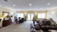This single-floor space functions as a combined lounge and dining area designed for comfort and casual gathering. The room features a textured ceiling with recessed lighting, which provides even, ambient illumination throughout. Warm beige walls contribute to an inviting atmosphere, complemented by wall sconces that add a soft, decorative glow on either side of the central French doors.

The French doors serve as a focal point along the back wall, flanked by large windows fitted with white blinds that allow generous natural light to fill the space. The patterned carpeting adds visual texture underfoot and ties the room's neutral color palette together.

The lounge area is anchored by rich, dark leather sofas arranged for comfortable seating and conversation. Nearby, a round dining table with traditional dark wood chairs offers a dedicated space for meals or activities. Potted tropical plants and a floral arrangement with sunflowers introduce natural elements, enhancing the welcoming ambiance.

A decorative ladder shelf along one wall provides storage and display space. Overall, the room balances functionality with a warm, residential feel, making it well-suited as a shared community or gathering space.