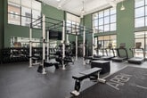 This two-story fitness center in Meridian, Idaho offers a well-equipped, thoughtfully designed workout facility spanning three dedicated exercise rooms across two floors. The main floor commands attention with soaring double-height ceilings, expansive floor-to-ceiling gridded windows that flood the space with natural light, and sage-green accent walls that lend a fresh, modern aesthetic. A striking mountain landscape mural serves as a dramatic focal point, reinforcing a nature-inspired design theme throughout. The main exercise area is outfitted with a full strength training zone featuring power racks, barbells, weight plates, and adjustable benches, alongside a dedicated cardio row featuring multiple treadmills, rowing machines (including RowERG rowers), ellipticals, stair climbers, and stationary bikes. Exposed ductwork and industrial pendant lighting contribute to the facility's contemporary character. A steel staircase leads to the upper level, which houses additional cardio equipment, medicine balls, resistance training stations, and open floor space for functional training. A separate, enclosed studio room on the main floor includes mirrored walls, a wall-mounted TV, dumbbell storage, and roller shades for privacy. Durable rubber flooring throughout ensures safety and performance across all zones.