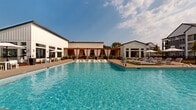 This single-story amenity complex is designed around a expansive, crystal-clear swimming pool that serves as the centerpiece of the outdoor environment. The architecture follows a modern farmhouse aesthetic, characterized by white vertical board-and-batten siding, dark-framed floor-to-ceiling windows, and clean architectural lines that create a cohesive and polished appearance throughout.

Surrounding the pool, a generously sized concrete deck is lined with cushioned lounge chairs and shaded umbrella stations, offering ample space for relaxation. A striking wood pergola structure extends along one side of the complex, draped with flowing white curtains that can be drawn for privacy or shade. Beneath the pergola, a turf-covered recreation zone houses a ping-pong table, while ceiling fans and string lights add comfort and ambiance for evening use.

An adjacent covered patio functions as an open-air lounge, furnished with light wood-framed seating, accent tables, and decorative pendant lighting. Large glass-panel doors and transom windows allow natural light to flood the interior lounge area, which features neutral tones, layered textiles, and contemporary furnishings. A glass-panel gate provides secure access to the pool area, complemented by manicured landscaping with shrubs and mulched garden beds throughout.