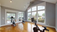 This single-floor fitness studio offers a welcoming and energizing environment designed for group cycling and active wellness activities. The space is defined by its striking vaulted ceiling, which soars overhead and contributes to an open, airy atmosphere. A ceiling fan is centrally mounted, providing comfortable airflow throughout sessions.

Warm hardwood flooring runs throughout the studio, offering both durability and an inviting aesthetic underfoot. The room is lined with expansive, floor-to-near-ceiling windows along multiple walls, allowing an abundance of natural light to fill the space and offering views of the surrounding greenery and outdoor landscape. These large windows give the studio an open, connected feel throughout the day.

Full-length framed wall mirrors are positioned along one side of the room, effectively doubling the perceived depth of the space while allowing participants to monitor their form during workouts. The walls are finished in a neutral gray tone, creating a clean and focused environment.

The studio is equipped with multiple Peloton spin bikes and additional cycling equipment, arranged to accommodate group classes or individual sessions. A set of white double doors provides a secondary entry point, while a glass-panel door connects to an adjacent area. An illuminated exit sign ensures clear safety egress. The overall design balances functional fitness infrastructure with a bright, polished aesthetic.