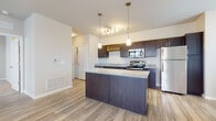 This single-floor unit is designed with a cohesive, modern aesthetic that emphasizes both functionality and style. The open-concept kitchen serves as a central feature, showcasing dark espresso cabinetry that contrasts sharply with the speckled granite countertops. A center island with matching cabinetry and granite provides additional prep and seating space, while pendant lighting above the island adds focused illumination. A built-in wine rack above the upper cabinets adds a distinctive design detail. The kitchen is fully equipped with stainless steel appliances, including a refrigerator, electric range, microwave, and dishwasher, all complemented by a double basin sink.

Wood-look luxury vinyl plank flooring extends through the kitchen and living areas, lending warmth and visual continuity. The living area is well-lit by natural light from multiple windows and a French door that leads to an outdoor area. The bedroom features plush carpeting and a double window with exterior views. The bathroom mirrors the kitchen's design language, with dark cabinetry, granite countertops, a large vanity mirror, and wood-look flooring. Overall, the unit maintains a clean, neutral palette with consistent finishes throughout.