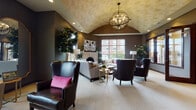 Located at 2911 Blue Aster Blvd in Sun Prairie, WI, this sophisticated single-floor space showcases refined design and carefully curated details throughout. The centerpiece of the space is a spacious lounge area distinguished by a dramatic barrel-vaulted ceiling with a textured gold finish and a striking orb chandelier that anchors the room. Deep charcoal walls create a rich, moody backdrop complemented by plush seating, including leather wingback chairs and neutral sofas accented with vibrant pink throw pillows. A white decorative fireplace serves as an elegant focal point, flanked by lush greenery and bold artwork. Oversized windows on multiple walls flood the living area with abundant natural light while offering open views of the surrounding neighborhood. The kitchen is outfitted with dark espresso cabinetry, gleaming stone countertops with dramatic veining, an arabesque tile backsplash, premium stainless appliances including a professional-grade range and a French door refrigerator, and rich hardwood flooring. A dedicated sitting area with floor-to-ceiling windows provides an intimate retreat bathed in natural light. Throughout the space, the cohesive use of dark tones, warm metallics, and thoughtful lighting creates an ambiance that is both inviting and distinctly upscale.