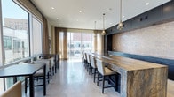 This contemporary single-floor space is designed with sophistication and functionality in mind, offering an inviting atmosphere ideal for gathering and entertaining. At the heart of the room is a commanding bar-height island clad in richly veined natural stone, complemented by an eye-catching mosaic tile backsplash and sleek dark cabinetry that extends along the back wall. Elegant pendant lights in a teardrop silhouette hang above the island, casting a warm glow over the cream upholstered bar seating arranged along its length. The polished concrete flooring reflects light beautifully, enhancing the open, airy feel of the space. Floor-to-ceiling windows dressed in sheer golden drapes flood the interior with natural light and frame dynamic urban views of the surrounding cityscape. Additional high-top tables and chairs are positioned near the windows, offering flexible seating arrangements for various uses. Recessed ceiling lighting throughout ensures the space remains well-lit at all times. The overall design blends industrial and refined elements seamlessly, creating a stylish, modern environment with a welcoming ambiance.
